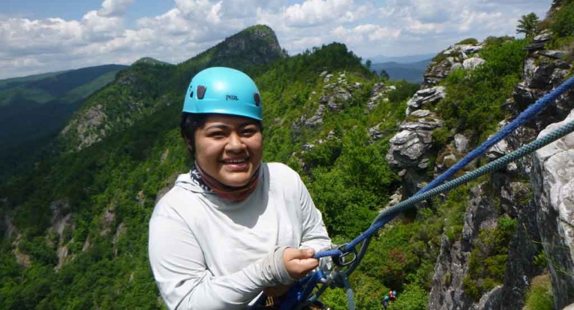 a high school student who is rock climbing smiles at the camera on an outward bound trip in north carolina